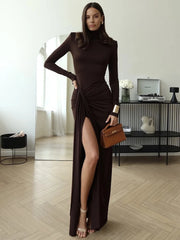 Woman in a long brown dress holding a brown handbag in a room with a modern interior.