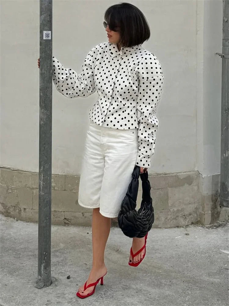 Woman wearing a polka dot blouse, white shorts, red shoes, and holding a black bag against a plain wall.