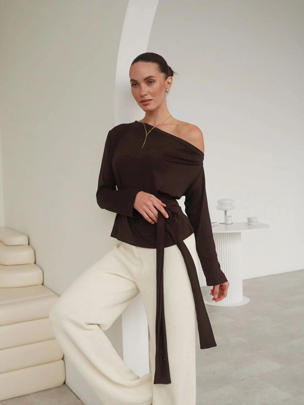 Woman wearing a brown off-shoulder top with a belt in a minimalistic room.