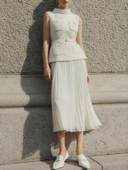 Person wearing a sleeveless beige top and pleated white skirt against a textured wall.