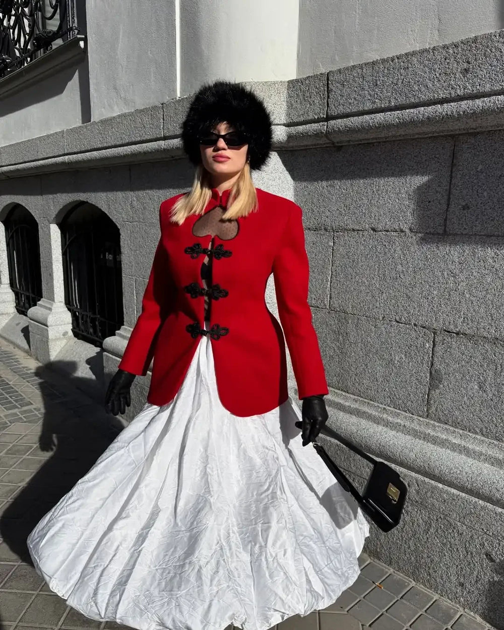Woman in a red coat and white dress standing against a stone wall.