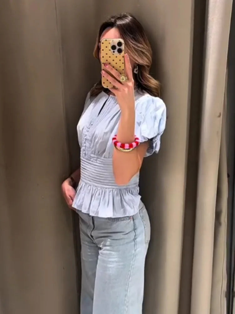 Woman taking a mirror selfie wearing a light blue top and jeans against a beige wall.