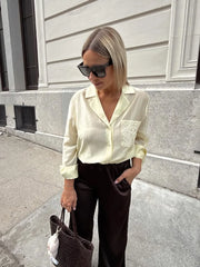 Woman wearing a light-colored blouse with a logo and dark pants, standing on a street.