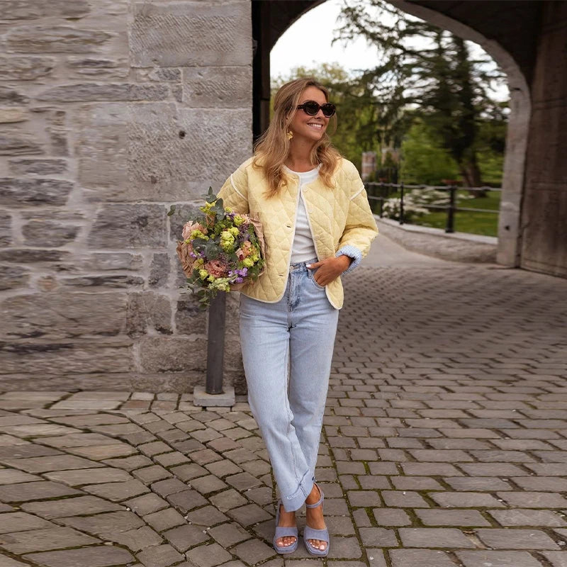 Woman in yellow jacket and light blue jeans holding flowers, standing on a stone pathway.