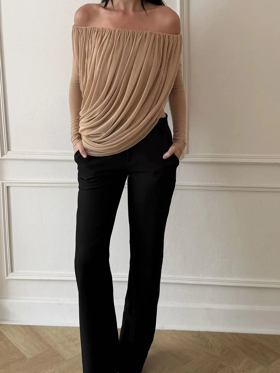 Person wearing a beige off-shoulder top and black pants against a white wall.