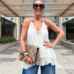 Woman in a white halter top and blue jeans holding a patterned clutch in an outdoor setting.