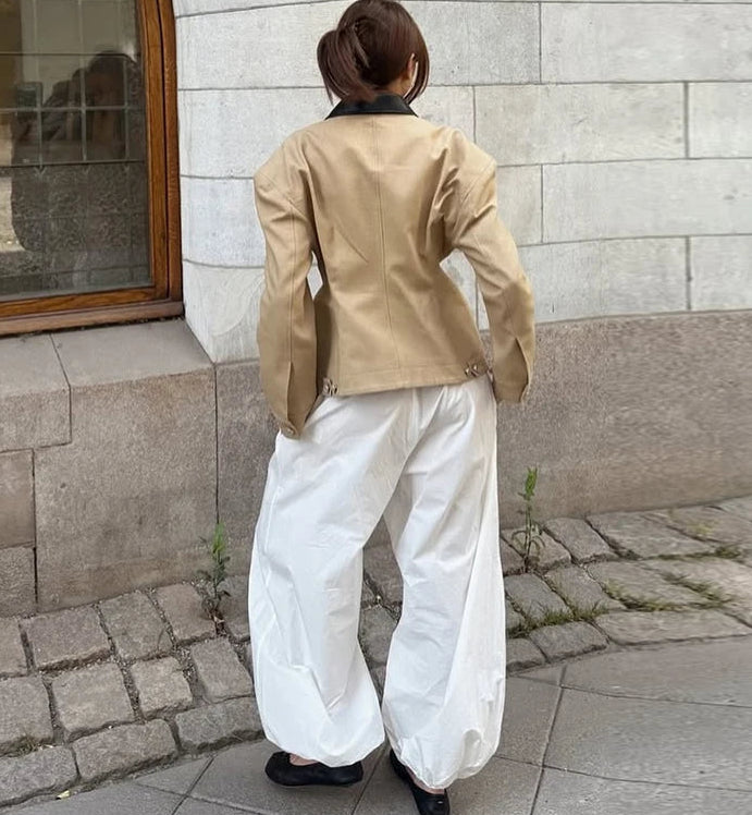 Person wearing a beige jacket and white pants standing against a stone wall.