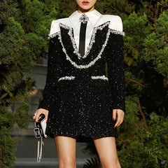 Person wearing a black sequin dress with a white shirt and black tie, holding a handbag, against a green foliage background.