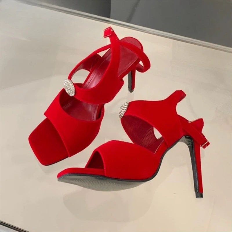 Pair of red stiletto heels with rhinestone embellishment and ankle strap on a reflective surface.