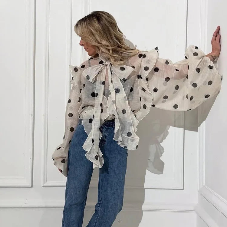 Woman wearing a polka dot blouse and jeans against a white paneled wall.