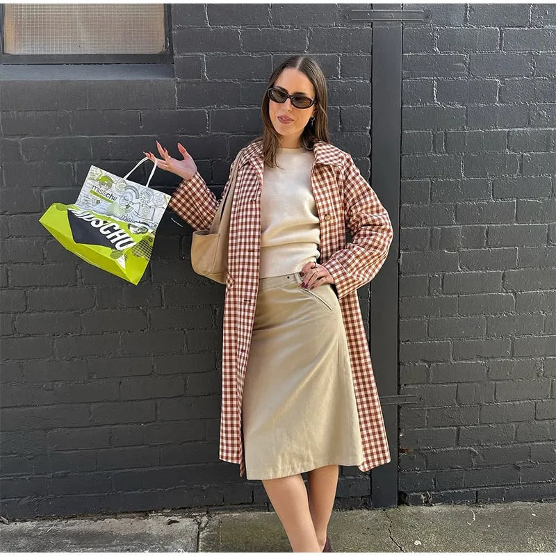 Woman in a plaid coat and beige skirt standing against a black brick wall, holding a shopping bag.
