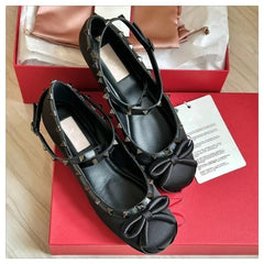 Black studded ballet flats with bow detail and ankle strap in box.