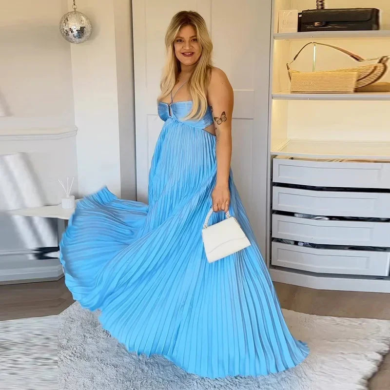 Woman in a blue pleated dress holding a white handbag in an indoor setting.