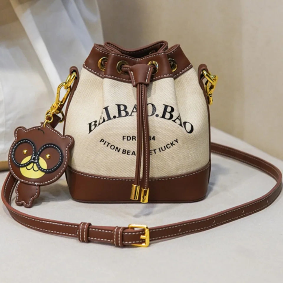 Beige canvas bucket bag with brown faux leather trim and bear charm.