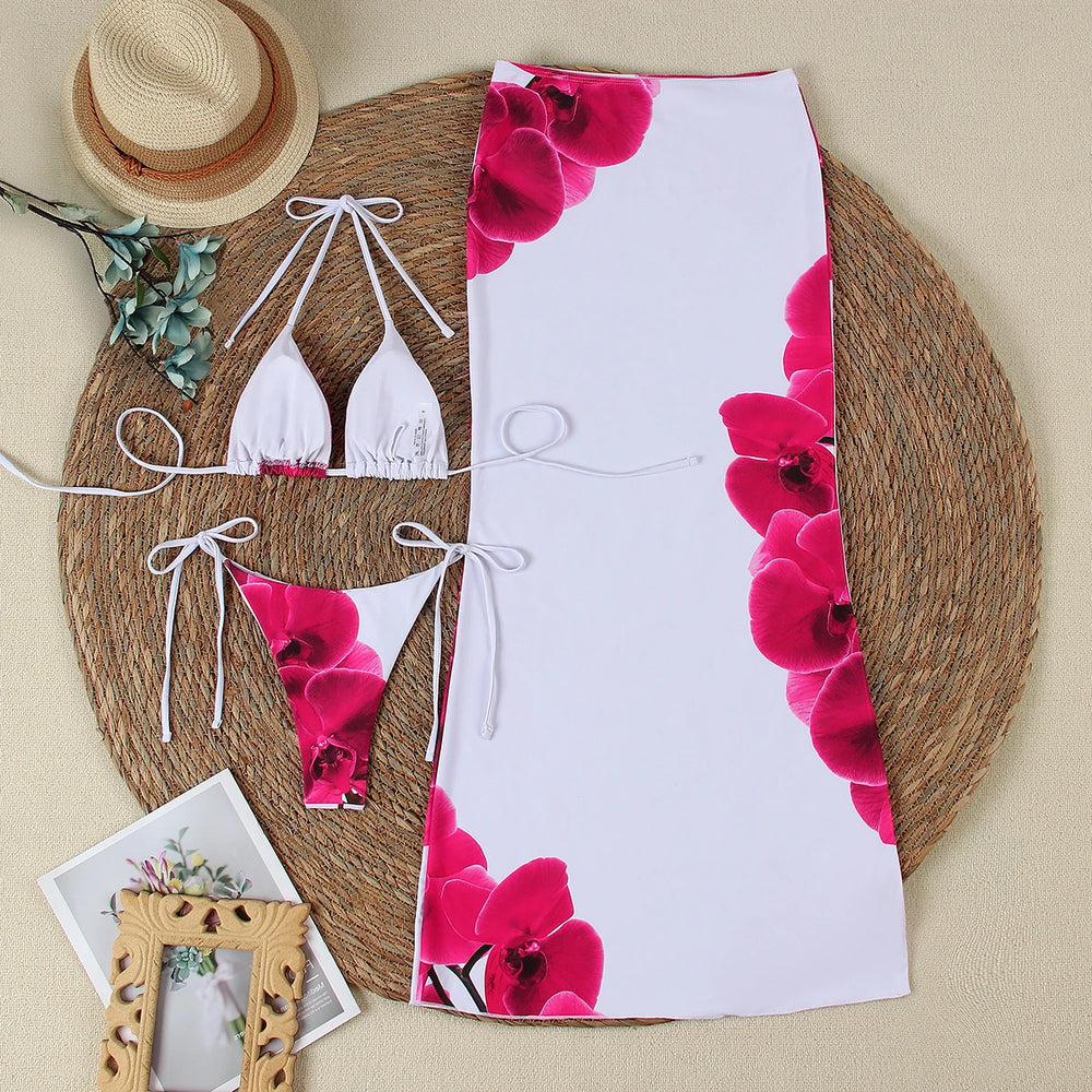 A flat lay of a white bikini top and bottom with pink orchid print, and a matching white maxi skirt with pink orchid print, arranged on a round woven mat.