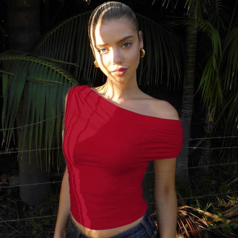 Woman wearing a red one-shoulder top standing in front of green foliage.