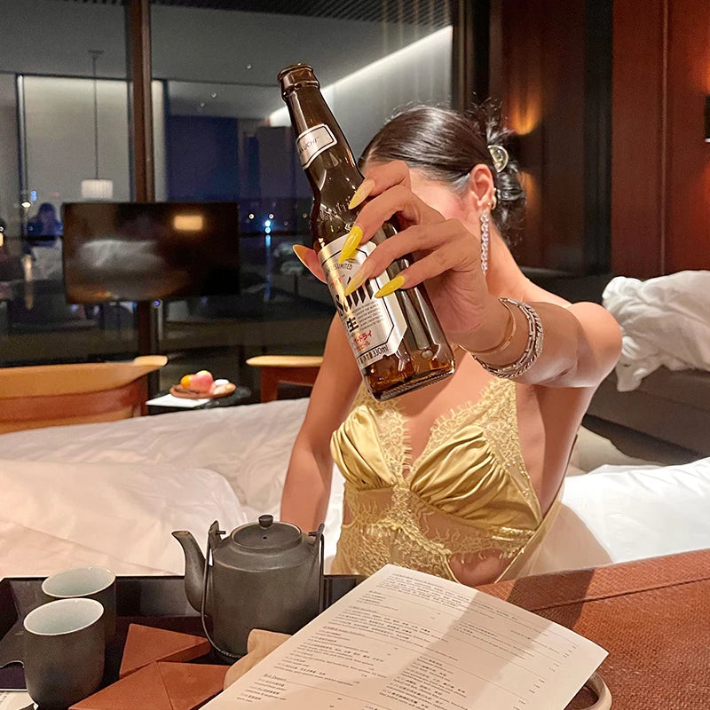 Woman in a gold dress holding a beer bottle in a room with a teapot and cups on a table.