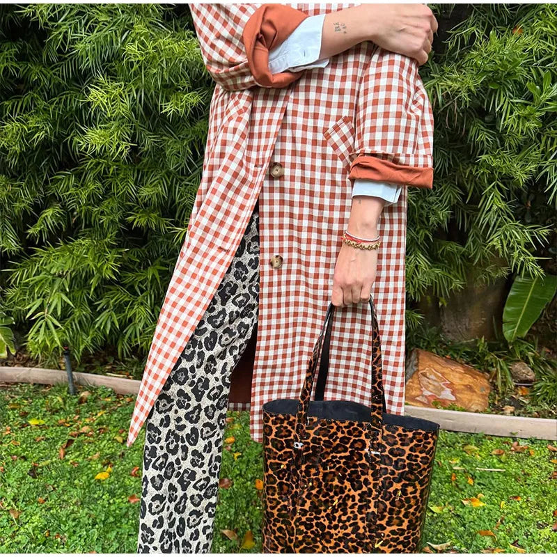 Person wearing a red and white checkered coat with leopard print pants and bag, standing in front of green foliage.