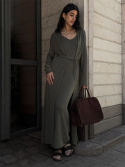 Woman in a long olive green dress holding a brown bag, standing against a building.