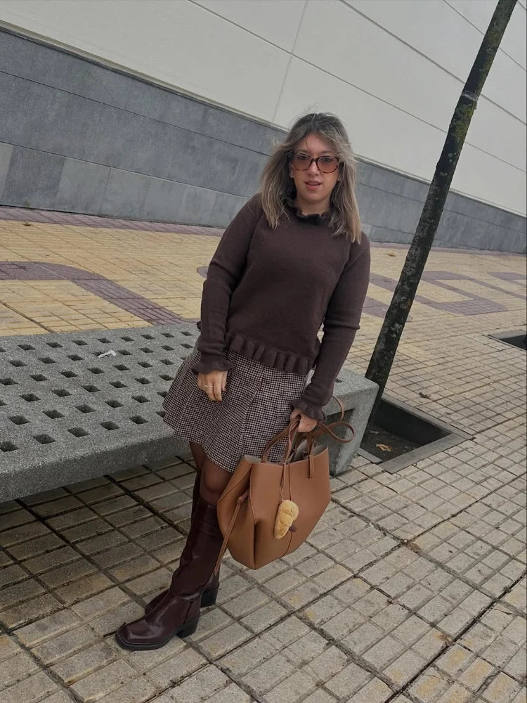 Woman in a brown sweater and skirt standing on a sidewalk with a brown handbag.