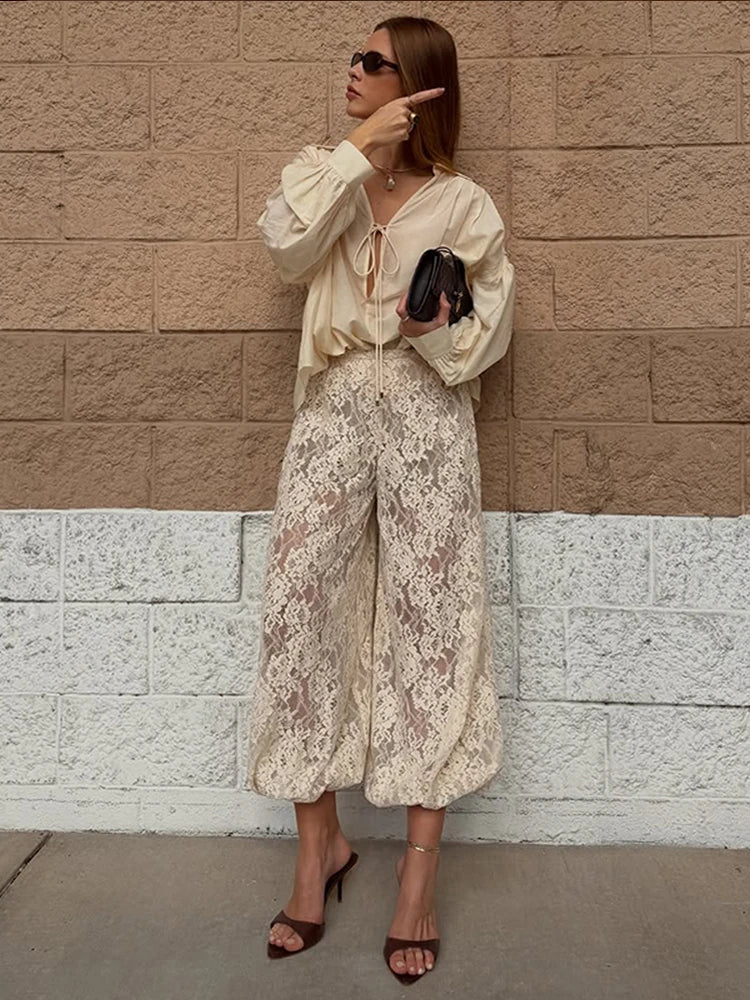 Woman in a stylish outfit with a beige blouse and lace pants against a brick wall.