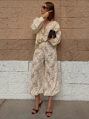 Woman in a stylish outfit with a beige blouse and lace pants against a brick wall.