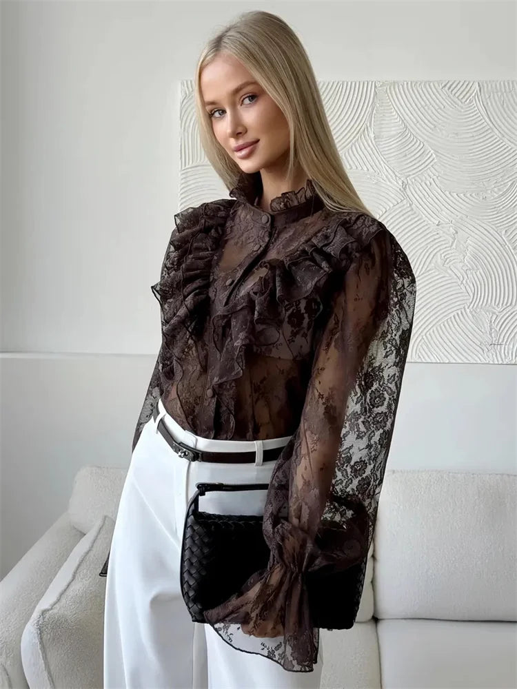 Woman wearing a sheer black lace blouse over a white top and pants, sitting on a couch.