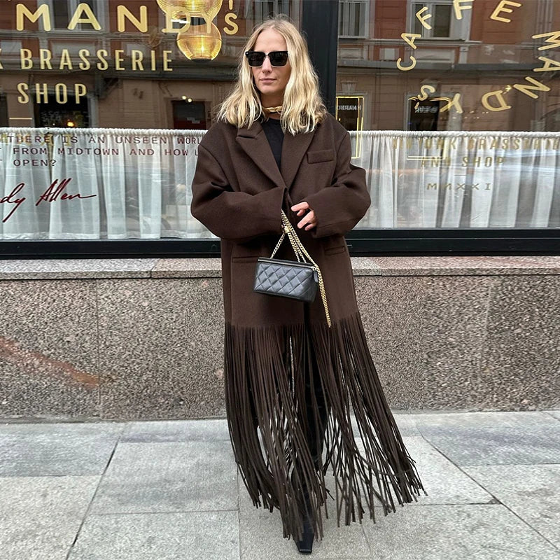 Woman wearing a brown fringed coat and sunglasses in front of a cafe.