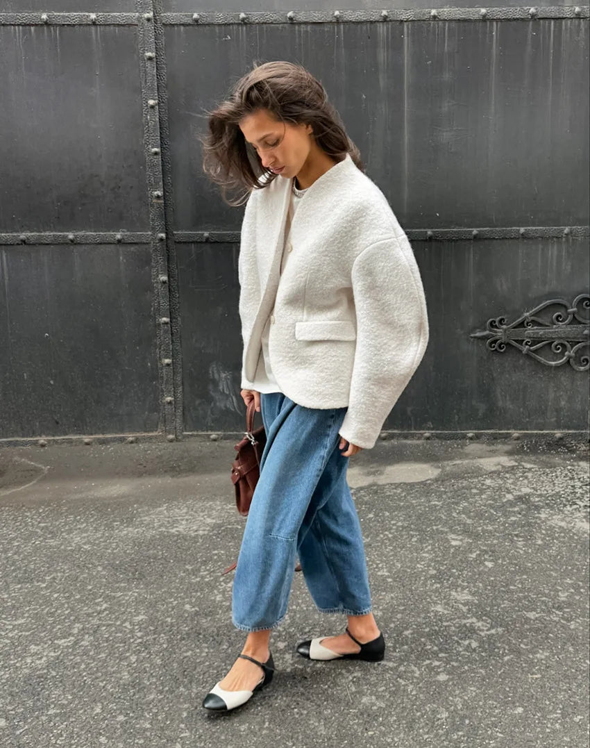 Woman wearing a white jacket and blue pants standing against a dark metal gate.
