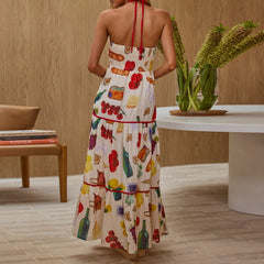 Woman wearing a colorful dress with a pattern of bottles, flowers, and fruits in a room with a wooden wall and a table.