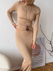 Person wearing a beige ribbed knit outfit with a pendant necklace against a white wall.