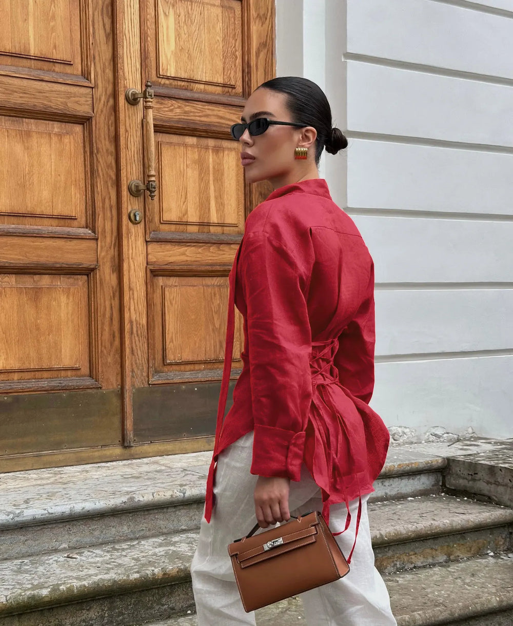 Woman in a red shirt and white pants holding a brown handbag, standing in front of a wooden door.