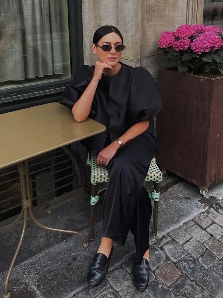 Person in black outfit sitting at an outdoor cafe table with flowers in the background