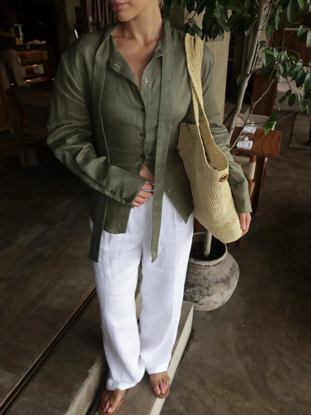 Person wearing a green shirt and white pants, holding a beige bag indoors.