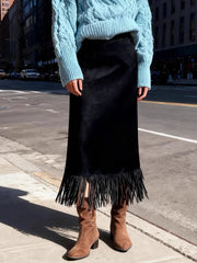 Person wearing a light blue cable knit sweater, black fringed skirt, and brown boots on a city street.