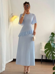 Woman wearing a light blue dress with ruffled sleeves in a room with a plant and lamp.