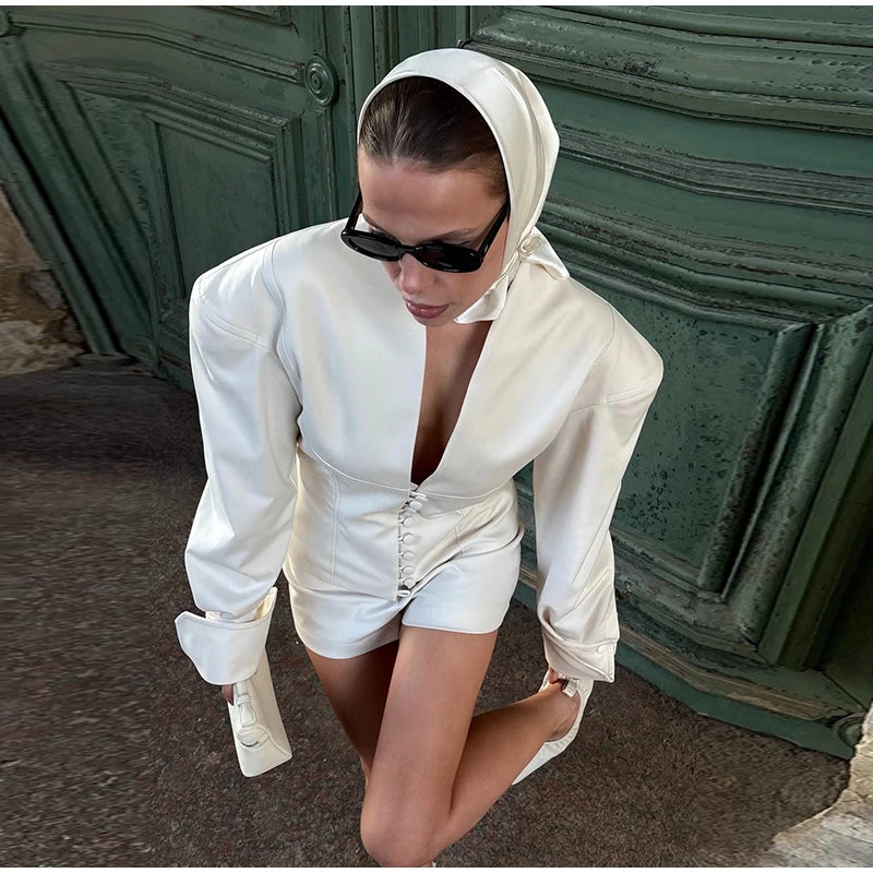Woman in a white outfit with a hood and sunglasses, posing in front of a green door.