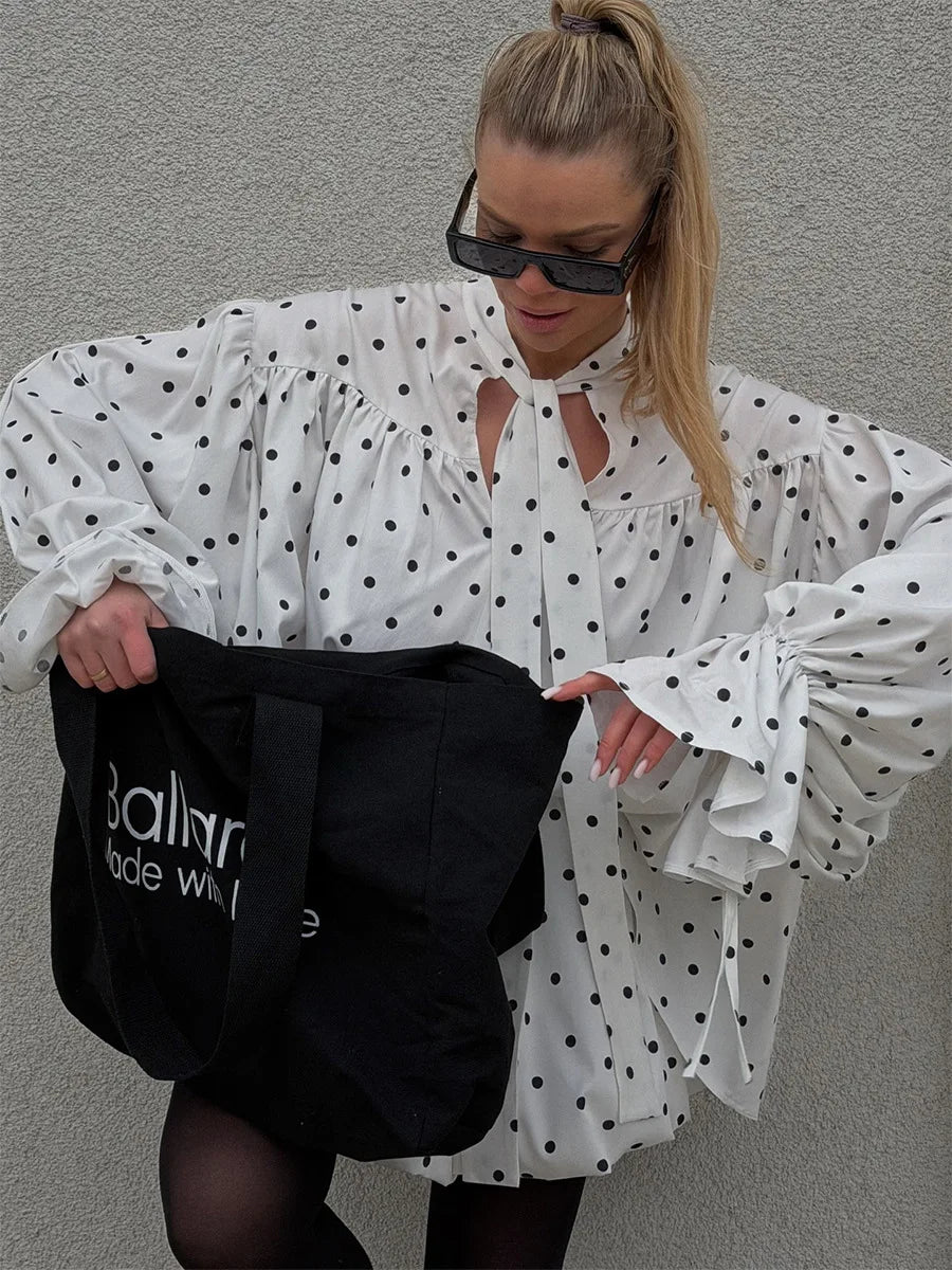 Woman wearing a white polka dot blouse holding a black bag with visible branding against a gray wall.