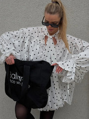 Woman wearing a white polka dot blouse holding a black bag with visible branding against a gray wall.