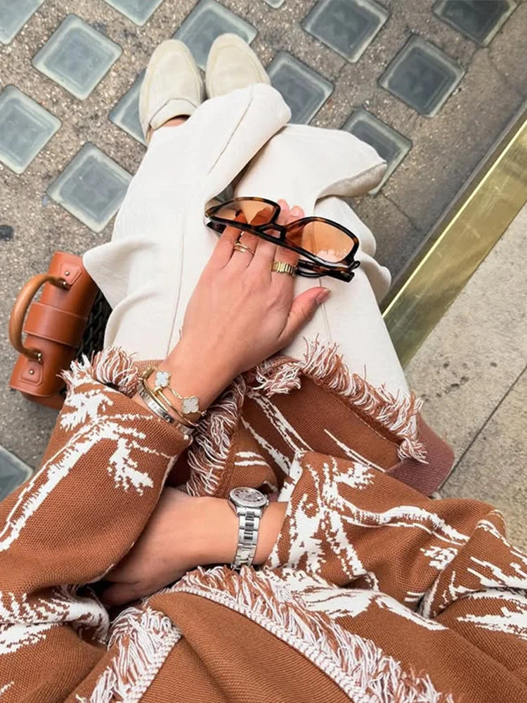 Person wrapped in a brown blanket with white patterns, holding sunglasses, on a tiled pavement.