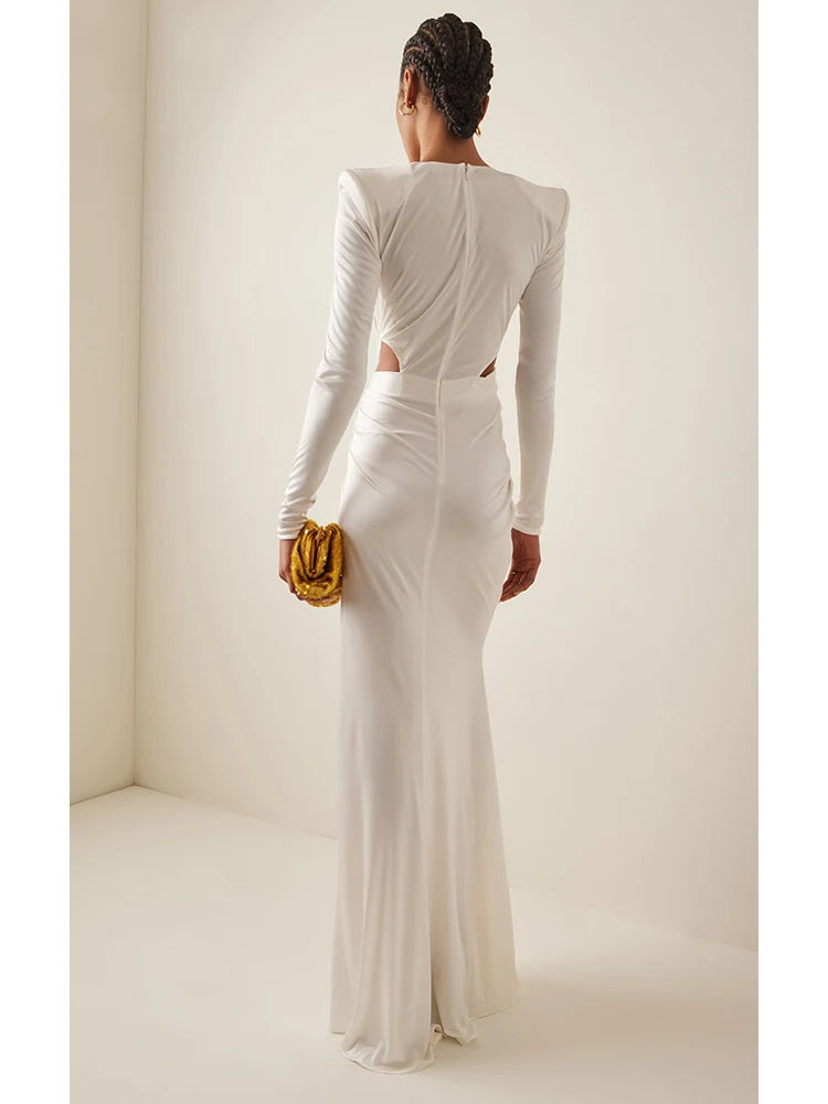 Woman wearing a white long-sleeve gown with cut-out design, holding a gold clutch against a plain background.
