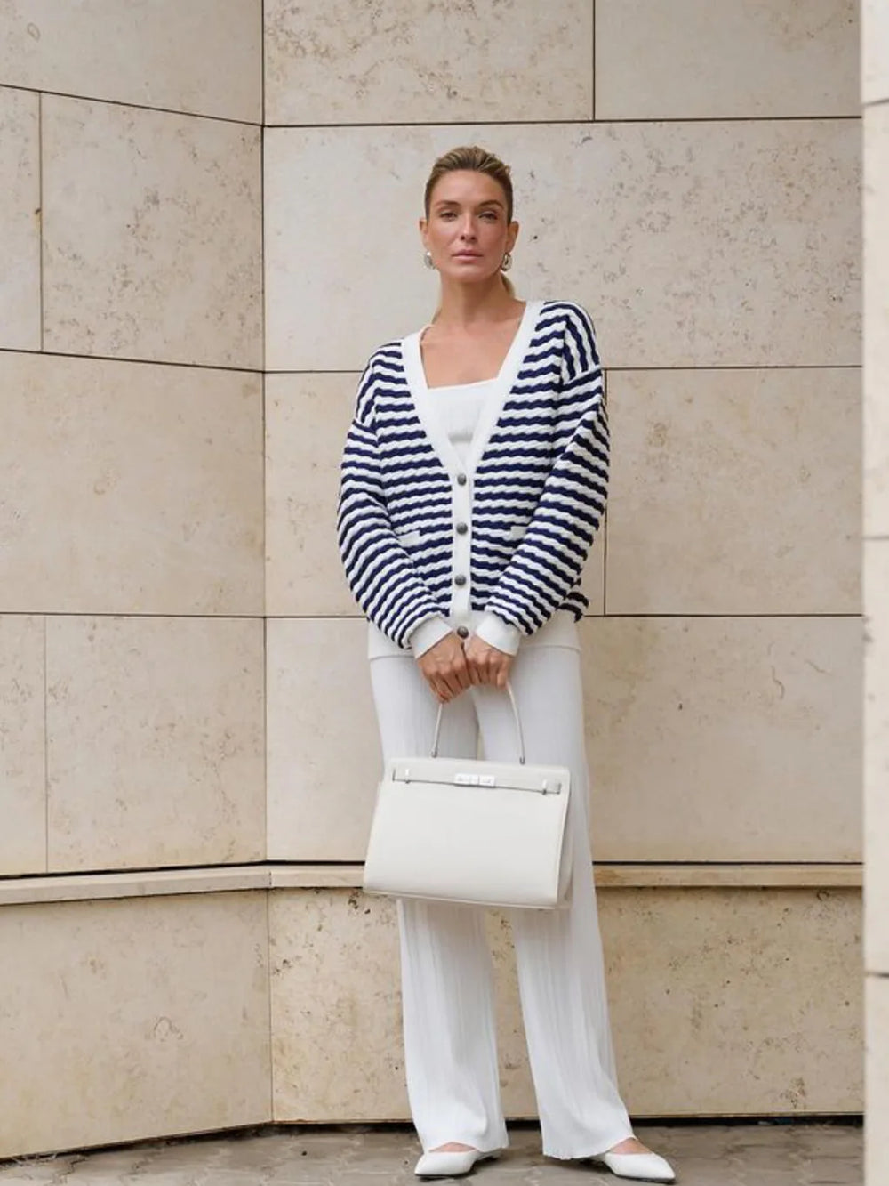 Woman wearing a striped cardigan, white shirt, and pants, holding a white handbag against a beige wall.
