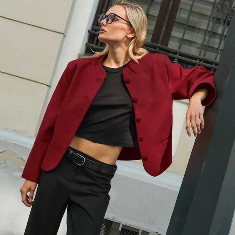 Woman wearing a red blazer over a black top and pants, standing against a building.