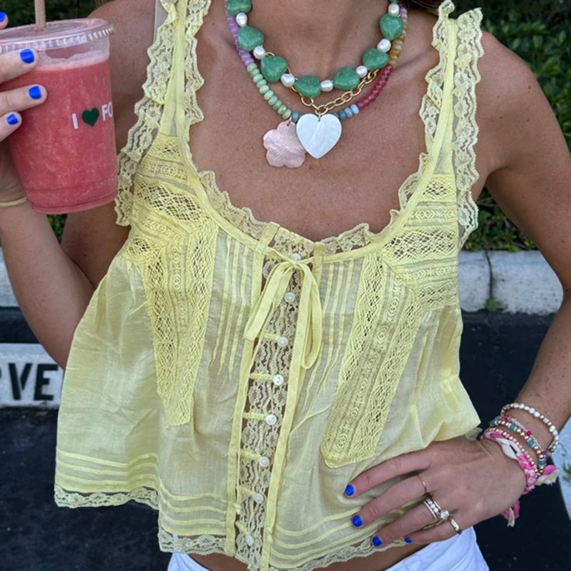 Person wearing a yellow lace top with a necklace and bracelets, holding a pink drink.