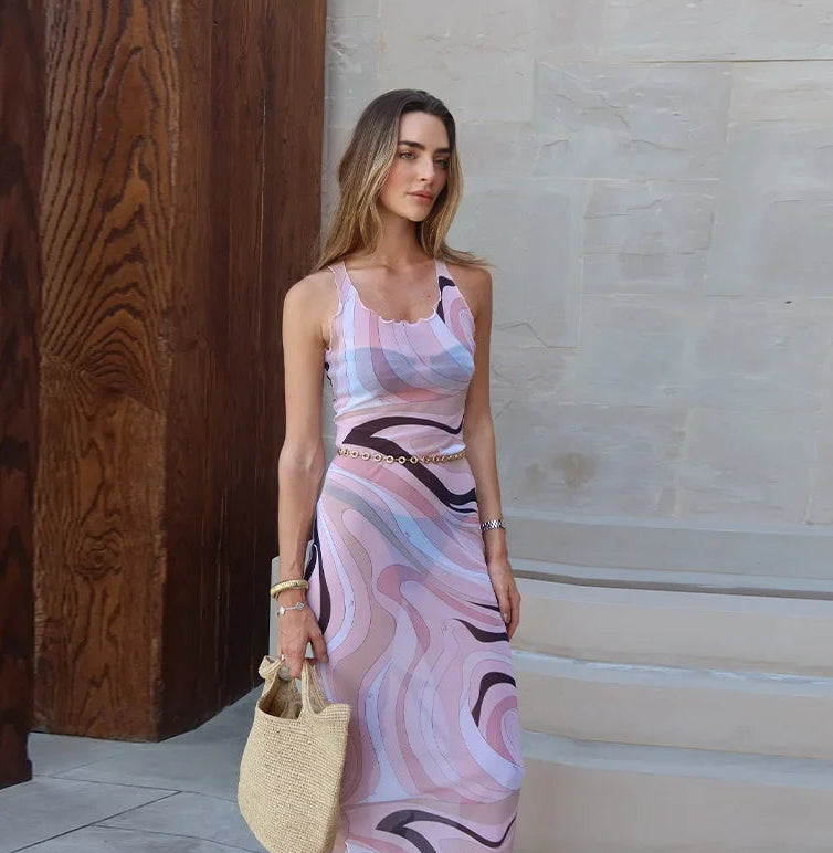 Woman in a patterned dress holding a straw bag against a stone wall.