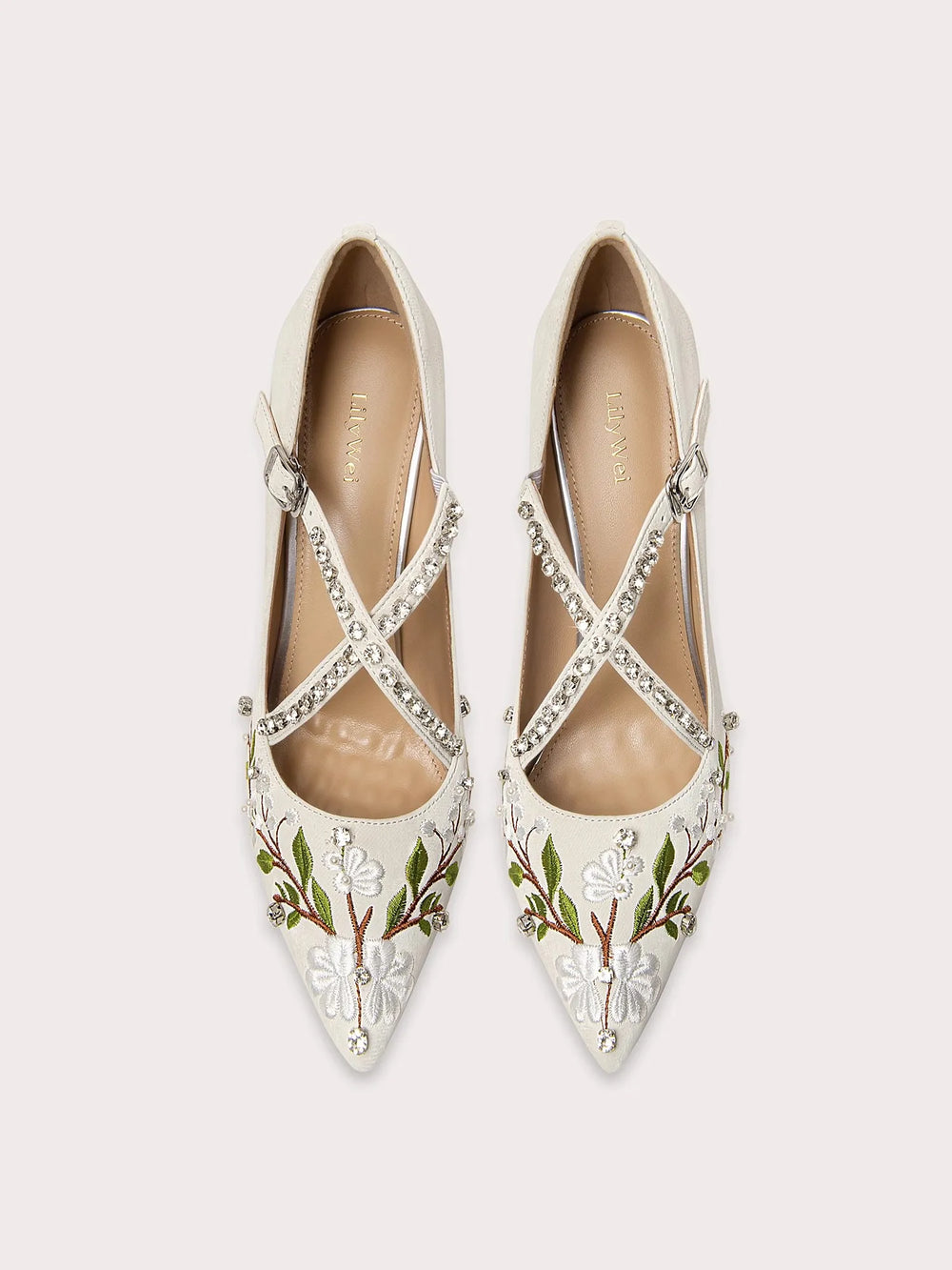 Pair of white pointed-toe shoes with floral embroidery and criss-cross straps on a light gray background.