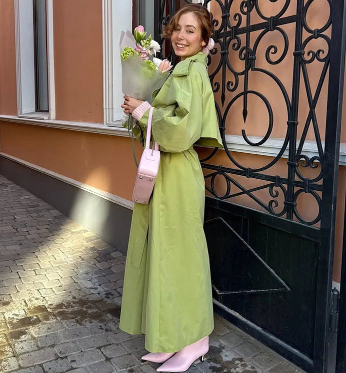 Woman in a green coat holding flowers and a pink bag standing in front of a building entrance.