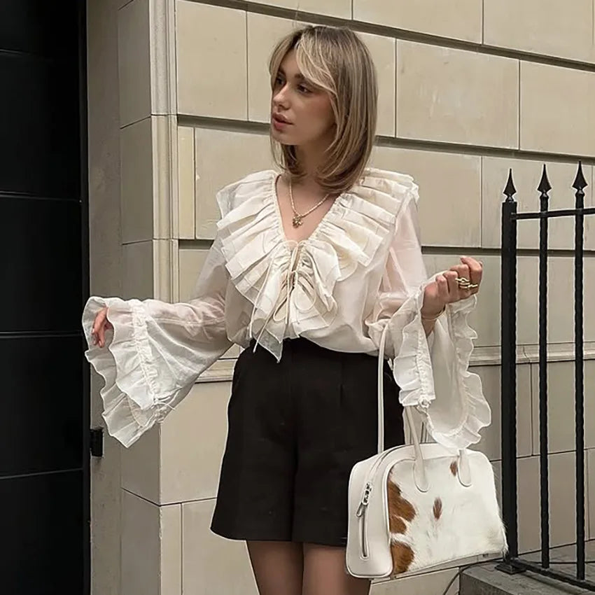 Woman wearing a white ruffled blouse and black shorts, holding a white handbag with brown spots, standing against a tiled wall.