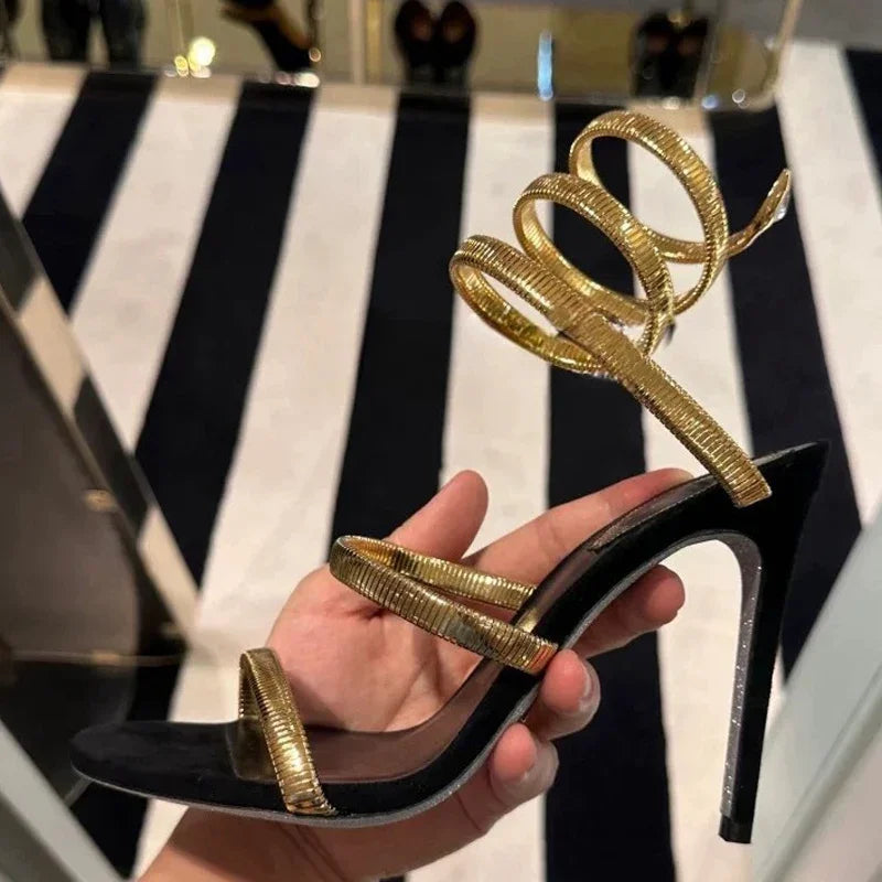 Black high-heeled sandal with gold straps held by a hand against a black and white striped background.
