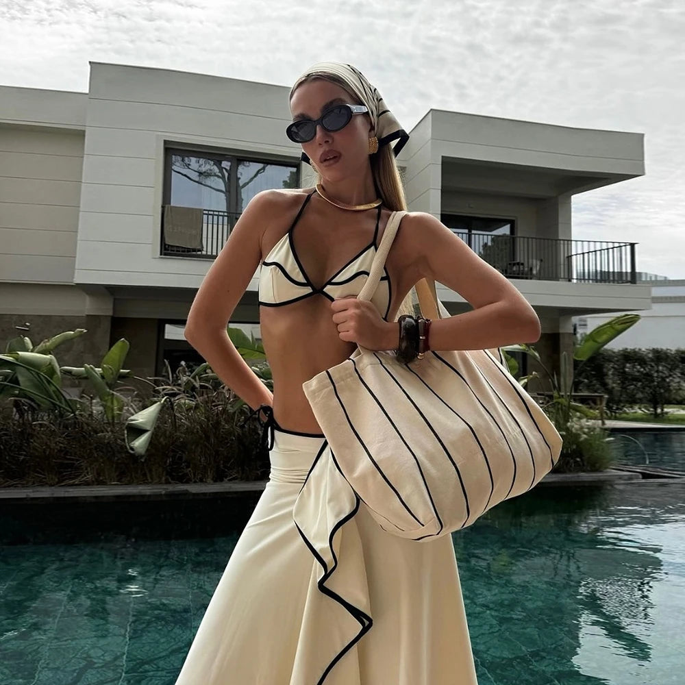 Woman wearing a white bikini with black contrast trim and a white ruffled maxi skirt with black trim, holding a striped tote bag by a swimming pool.